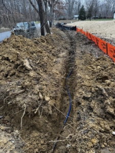 A long trench with a blue utility pipe and orange safety fencing, showing excavation work by Richardson Ridge Construction in Bloomington, IN.