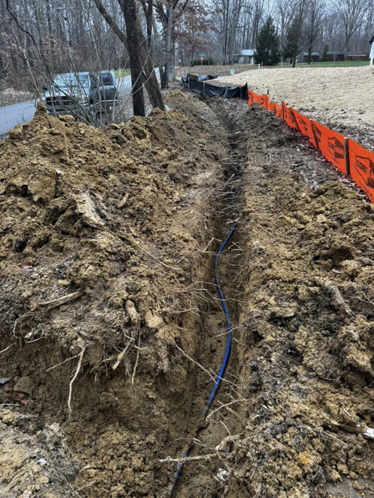 A long trench with a blue utility pipe and orange safety fencing, showing excavation work by Richardson Ridge Construction in Bloomington, IN.