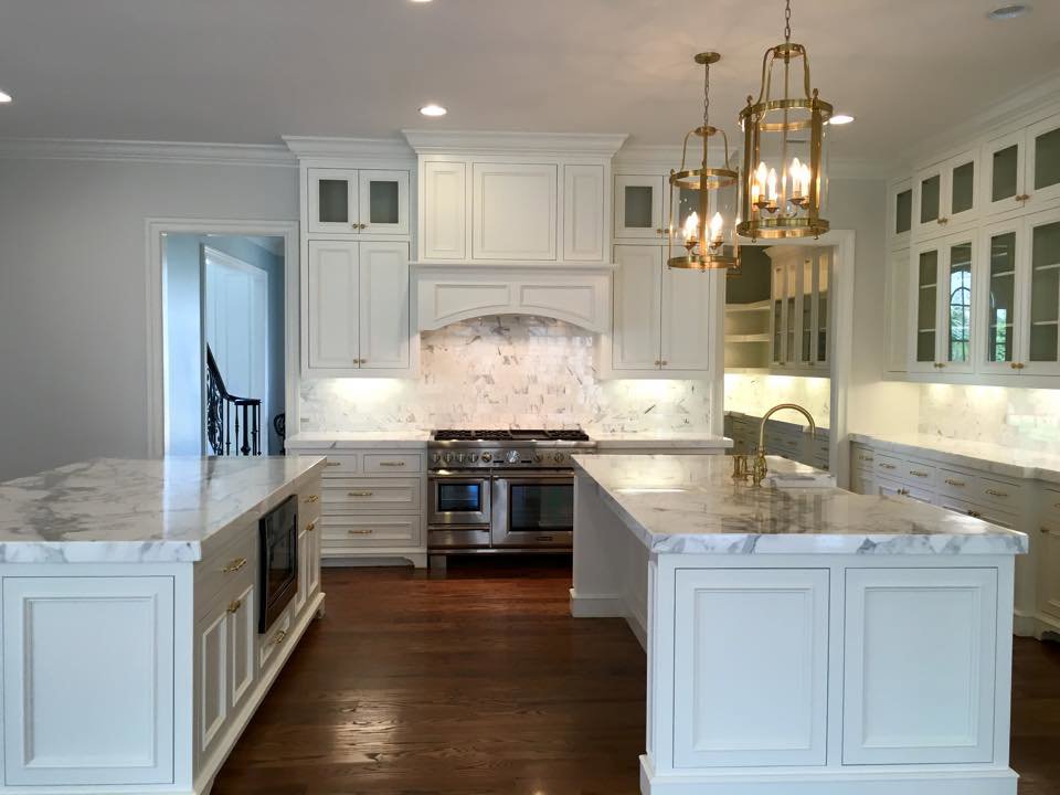 A luxurious white kitchen remodel featuring two large islands with marble countertops and elegant lighting by Pro Finishes NC, LLC in Raleigh, NC.