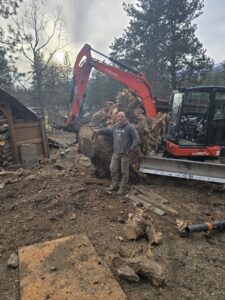 A man standing next to a large tree root ball with an excavator, showing stump removal by PJ's Tree Service in Missoula, MT.