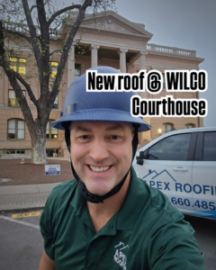 A man in a hard hat and APEX Roofing shirt in front of the Wilco Courthouse, indicating a new roof job by APEX Roofing in Georgetown, TX.