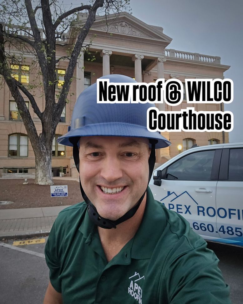 A man in a hard hat and APEX Roofing shirt in front of the Wilco Courthouse, indicating a new roof job by APEX Roofing in Georgetown, TX.