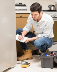 A man inspecting a refrigerator in a kitchen with a multimeter and toolbox, representing services by Hire A Hubby in Fresno, CA.