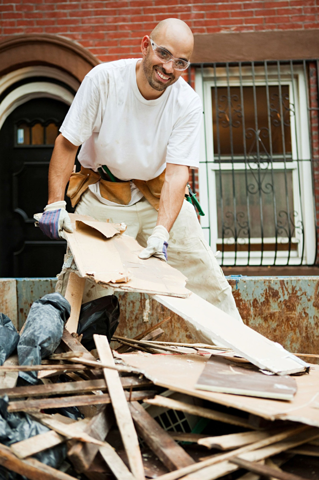 A smiling man in safety glasses loads cardboard into a large dumpster filled with debris, demonstrating junk removal services by Haul Gone 757 in Chesapeake, VA.