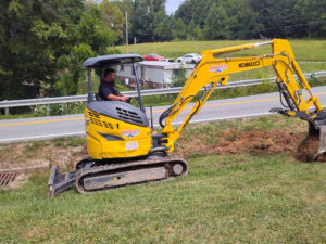 A man operating a mini excavator, digging a trench next to a road for Huntington Home Repair in Huntington, WV.