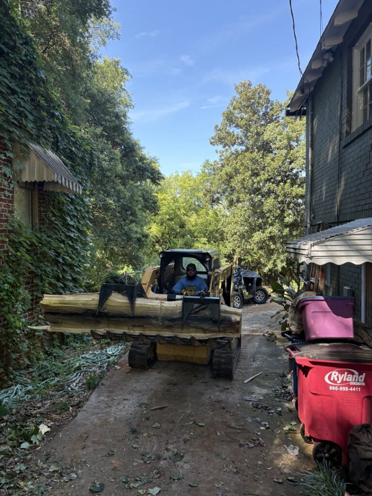 A man operating a compact track loader with a log grapple, moving a large log for Knotty Branches Tree Service in Macon, GA.