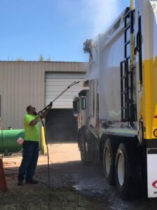 A man in a yellow shirt pressure washing a white garbage truck, maintaining equipment for Liberty Disposal, Inc. in Tucson, AZ.