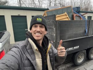 A man giving a thumbs-up next to a trailer loaded with furniture and junk at a storage facility, by Hometown Hauling 802 in Milton, VT.