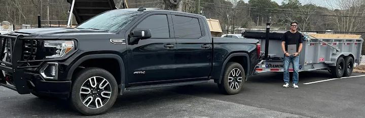 A man stands proudly next to a black pickup truck towing an empty dump trailer, ready for junk removal services by Lewis Outdoor Services LLC in Augusta, GA.