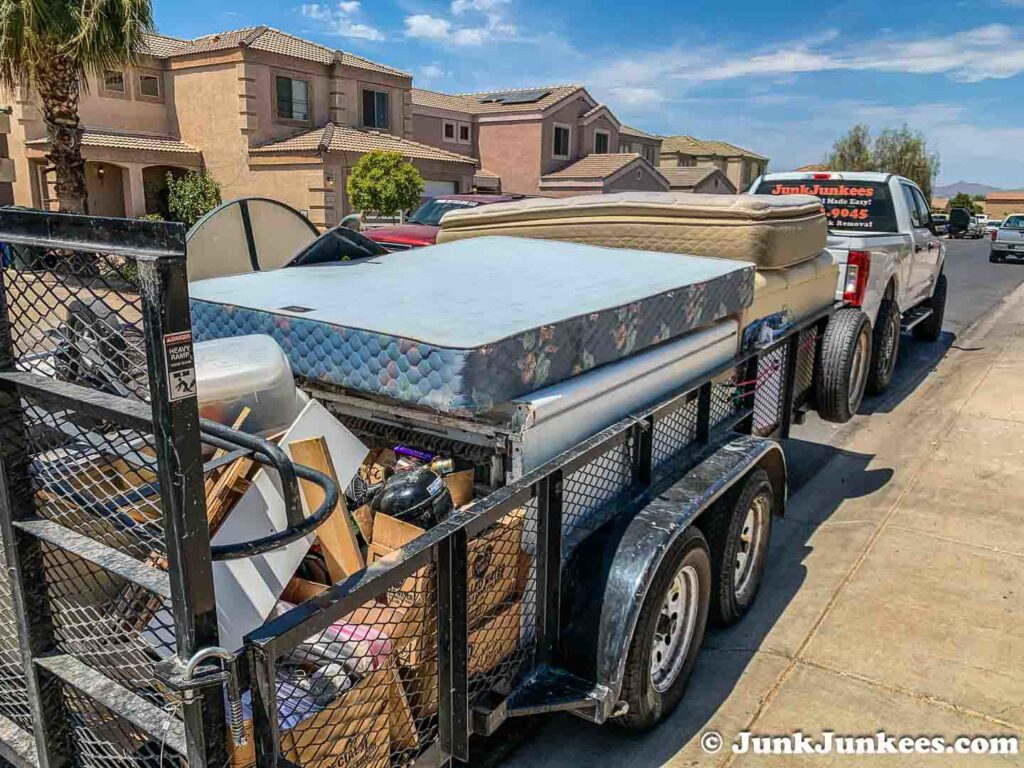 A Junk Junkees truck and trailer loaded with mattresses and household junk on a residential street in Peoria, AZ.