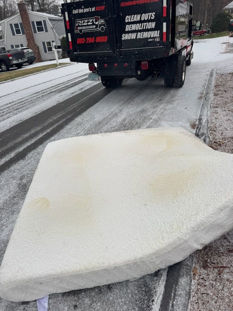 A mattress placed on the side of a snowy road with a Rizzo Junk Removal truck in the background, ready for pickup in Nashua, NH.