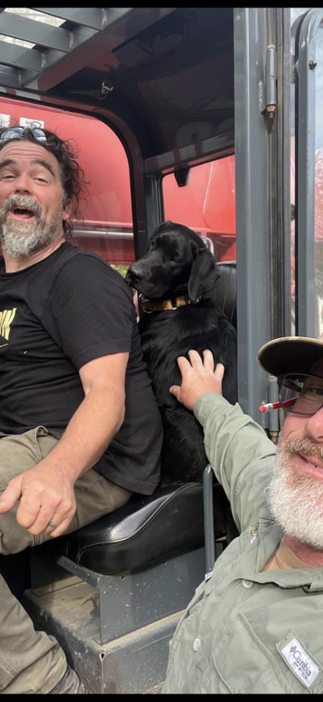 Two men and a dog in the cab of heavy machinery, likely on a job site for J & J Custom Builders LLC in Milford, CT.