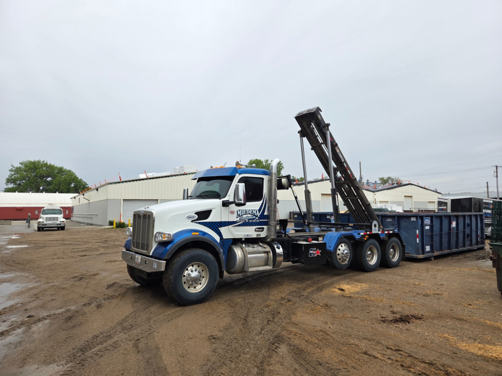 A Miedema Sanitation Inc of Huron SD roll-off truck with a blue dumpster on a dirt lot in Mitchell, SD.