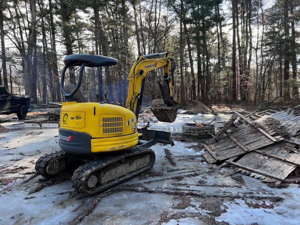 A yellow mini excavator clearing wooden debris from a snowy ground for Getting Trashed Dumpster Rentals & Excavation in Assonet, MA.
