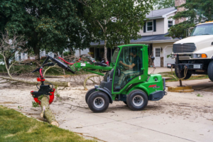 A green mini-loader with a grapple attachment clears cut tree branches from a residential street for Canopy Cops Tree Service LLC in Appleton, WI.