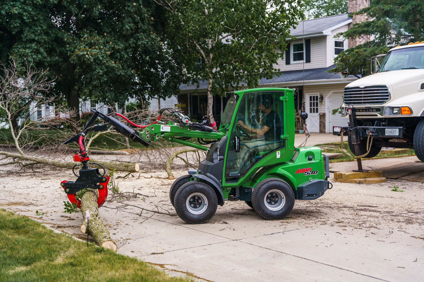 A green mini-loader with a grapple attachment clears cut tree branches from a residential street for Canopy Cops Tree Service LLC in Appleton, WI.