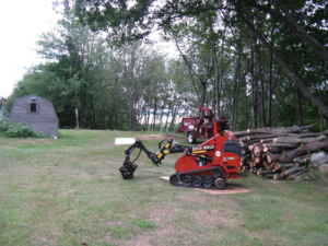 A mini skid steer with a grapple attachment next to a pile of cut logs by Fagan Tree Service in Brunswick, ME.