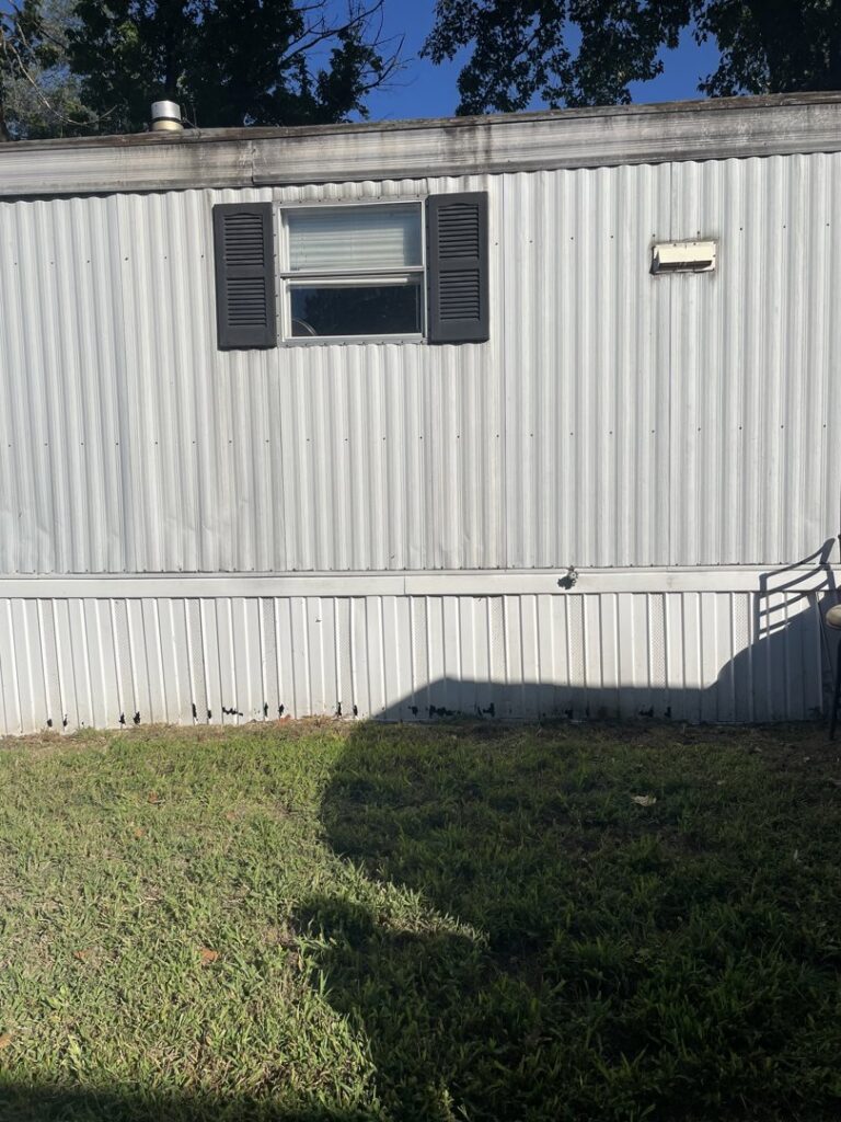 The exterior side of a mobile home with some dirt and debris at the base, indicating property preservation services by Little Tykes Property Preservation in Independence, MO.