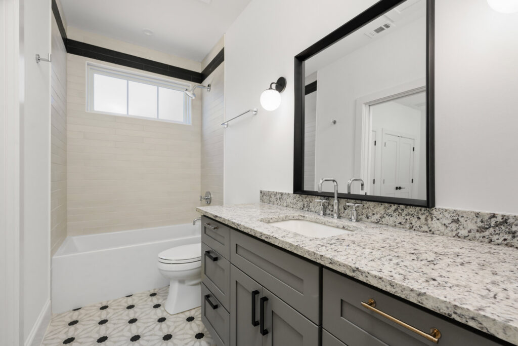A modern bathroom featuring a grey vanity, granite countertop, and a black and white patterned floor by Stone Acorn Builders in Bellaire, TX.