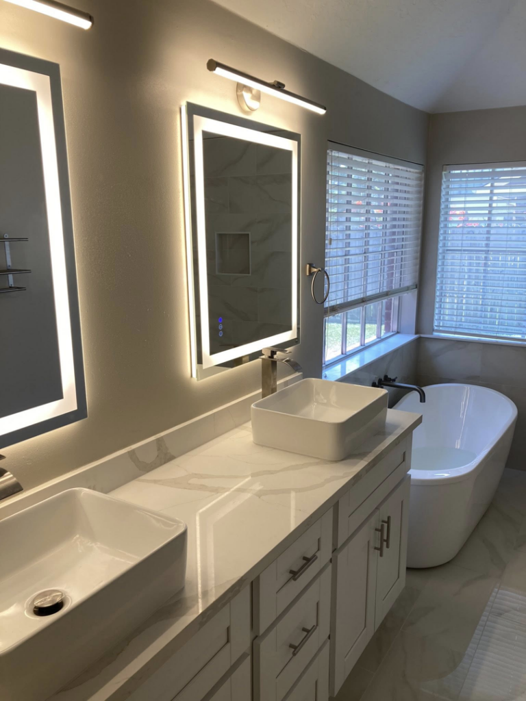 A modern bathroom renovation featuring a double vanity with lighted mirrors and a freestanding tub by J.C.Improvement in Katy, TX.
