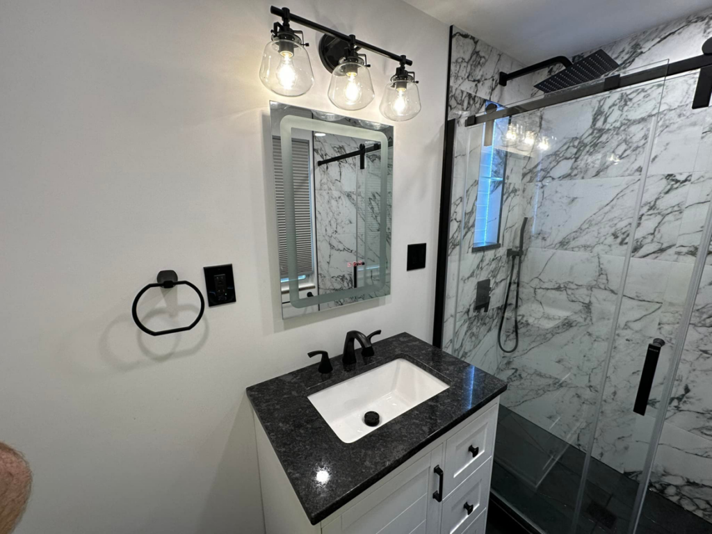 A modern bathroom vanity with black granite countertop, illuminated mirror, and shower by Tap & Tile Inc. in East Pittsburgh, PA.