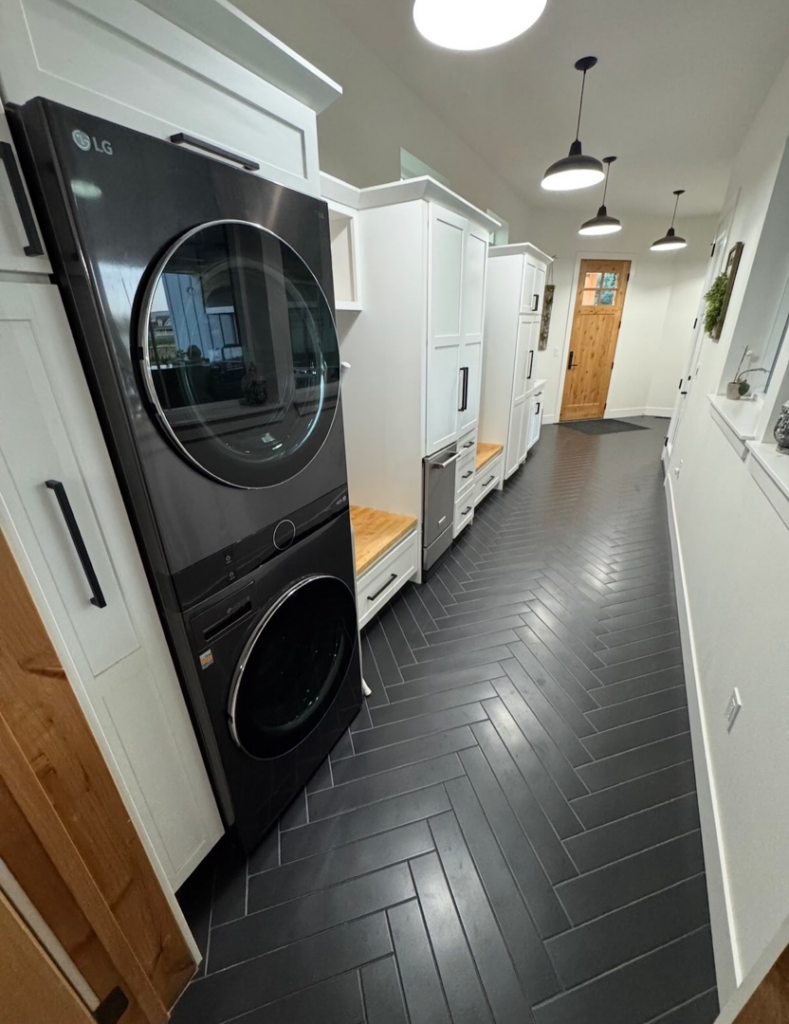 Newly installed modern laundry room with stacked washer/dryer, dark herringbone tile, and white cabinets by Fisher Construction, LLC in Selah, WA.