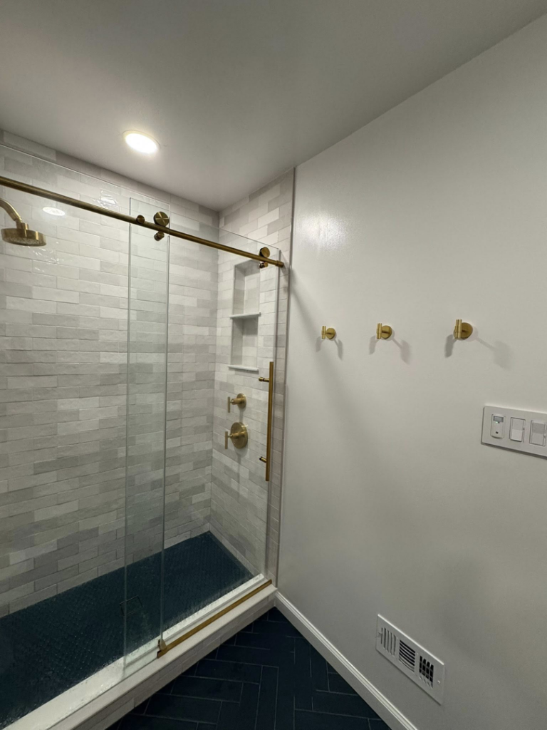 A modern shower with white subway tiles, dark blue floor tiles, gold fixtures, and a glass sliding door by NC Home Remodeling and Custom Cabinetry in Alexandria, VA.