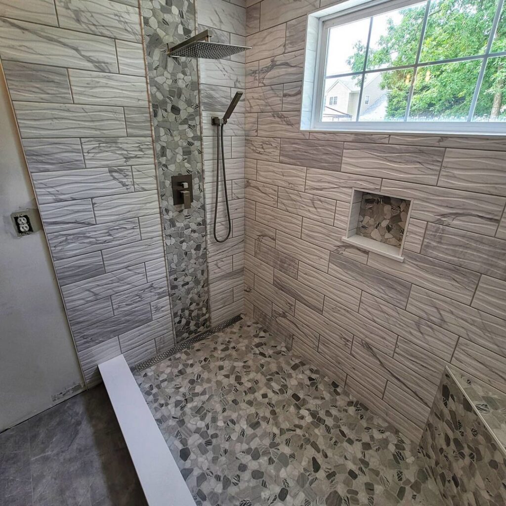 Modern shower with grey wavy tiles, a decorative pebble accent strip, and a rain shower head, installed by Supreme Tile & Flooring in Charlotte, NC.
