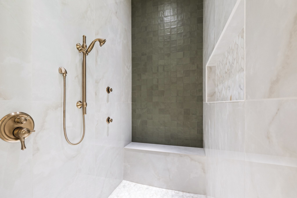 A modern shower installation featuring white marble tiles, a dark green accent wall, and bronze fixtures by Dugas Construction, LLC in Lafayette, LA.