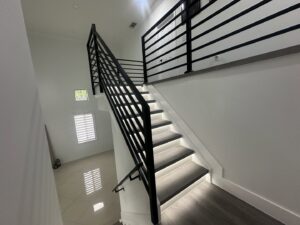 A modern staircase with sleek black metal railings and light wood-look treads featuring integrated LED lighting by SD Masters in Miami, FL.