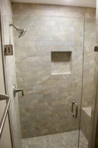 A modern walk-in shower with light rectangular tiles, a glass door, and a built-in niche, part of a bathroom remodel by Custer Remodeling in Plano, TX.