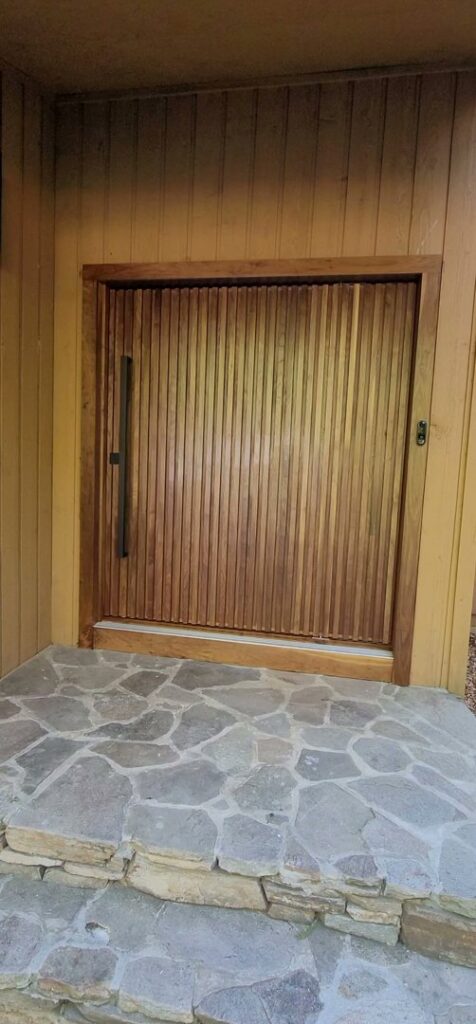 A newly installed modern wooden front door with vertical slats and a long black handle by M J Services Home Remodeling in Gaithersburg, MD.