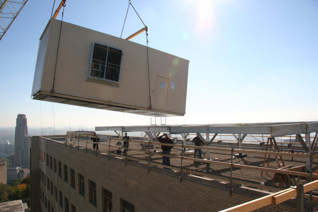 A large modular unit being lifted by a crane onto a building rooftop with workers, a construction project by McKamish, Inc. in Pittsburgh, PA