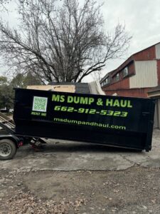 An MS Dump & Haul branded dumpster, partially filled with junk, parked on a residential driveway in Hernando, MS.