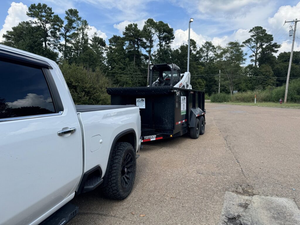 An MS Dump & Haul truck transporting a dumpster trailer with a skid steer loader on top in Hernando, MS.