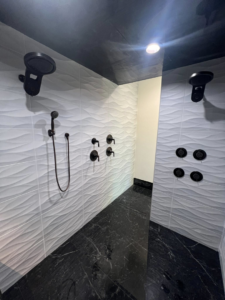 A spacious walk-in shower with multiple shower heads, textured wall tiles, and black marble floor by Patriot Tile LLC in Skiatook, OK.