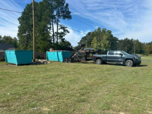 A truck with a trailer and two blue dumpsters on a grassy field by Site Boss Dumpster Rentals in Raeford, NC.