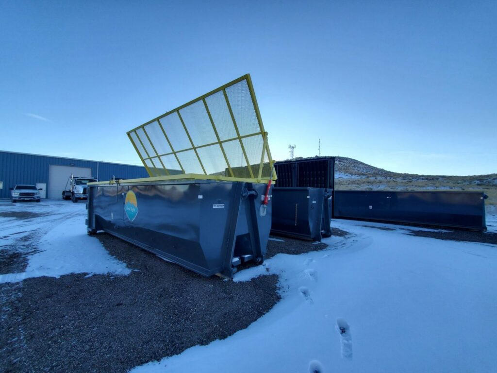 Multiple roll-off dumpsters, one with an open safety net, stored by First Light Services Llc in Casper, WY.