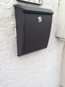A new black mailbox with an ornate design, securely installed on a textured white wall by AON Handyman in Wichita, KS.