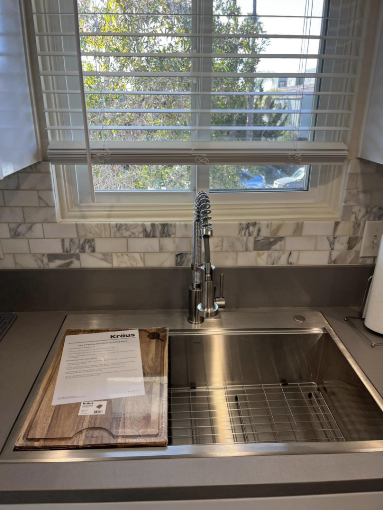 A newly installed stainless steel kitchen sink with a modern faucet and marble backsplash by Pauda Handyman in San Diego, CA.