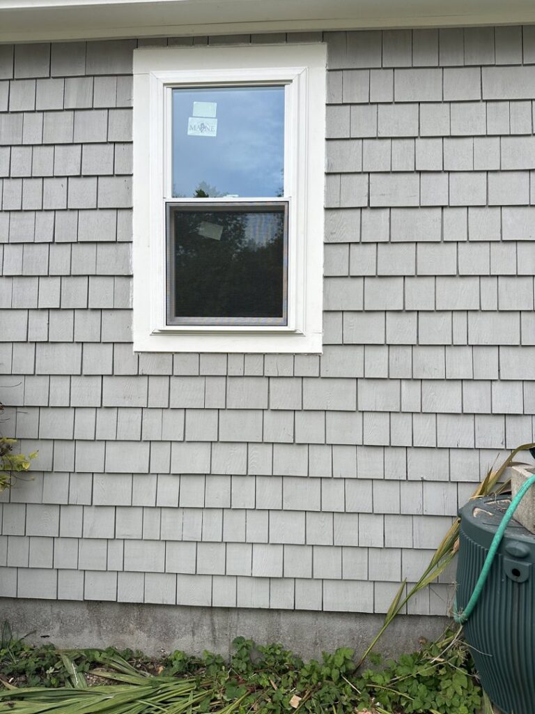 A newly installed window with a 'MAINE' sticker on a shingled exterior wall by Woodbine Repair & Remodeling in Jamestown, RI.
