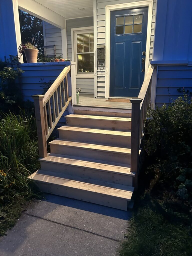 A newly constructed wooden porch with stairs and railings leading to a blue front door by Blackcap Handyman in Portland, ME.