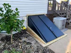 A newly installed black basement egress window well cover next to a house, installed by TCM Waterproofing, LLC in Canton, OH.