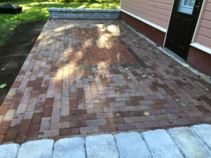 A newly installed brick paver patio with a small retaining wall next to a house by Senn II Excavating in Montague, MA.