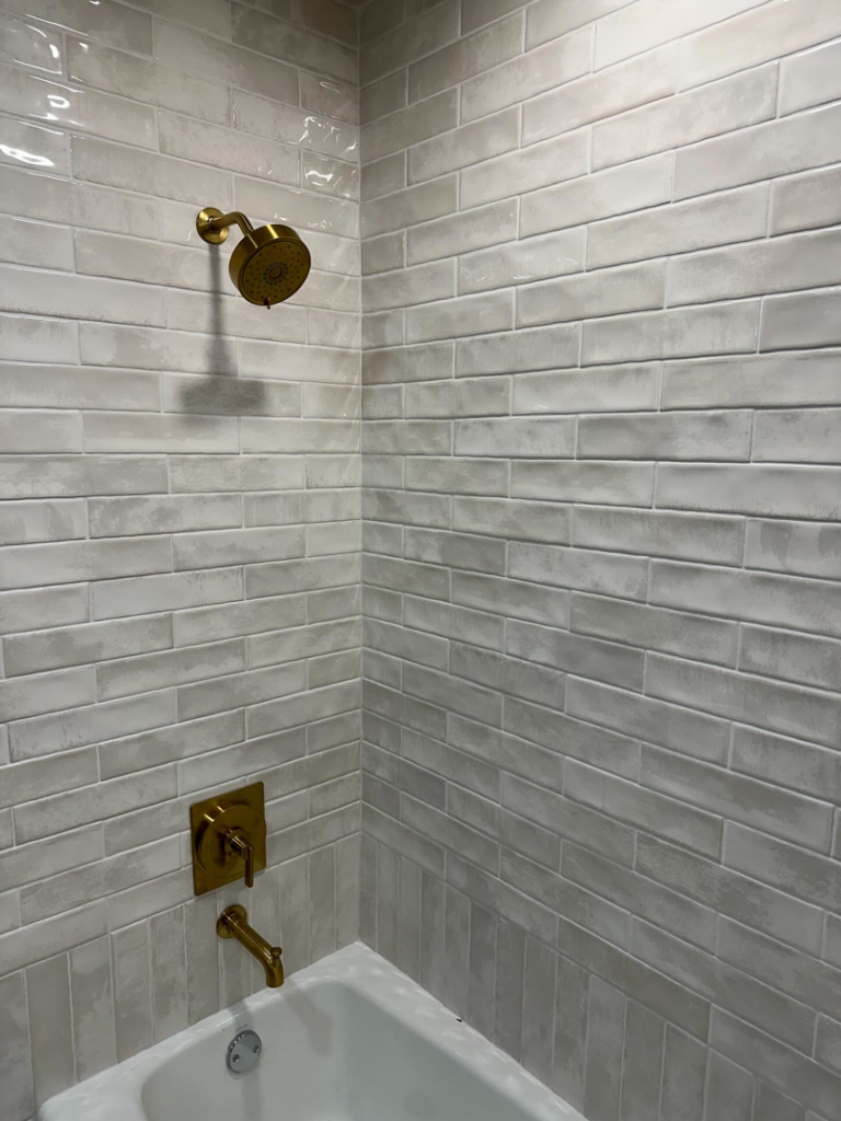 Newly installed gold shower head and faucet in a tiled shower by Le Roux Homes in Enid, OK.