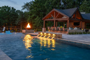 Night view of a custom pool with fire features and an outdoor pavilion by Elevate Pools in O'Fallon, MO.