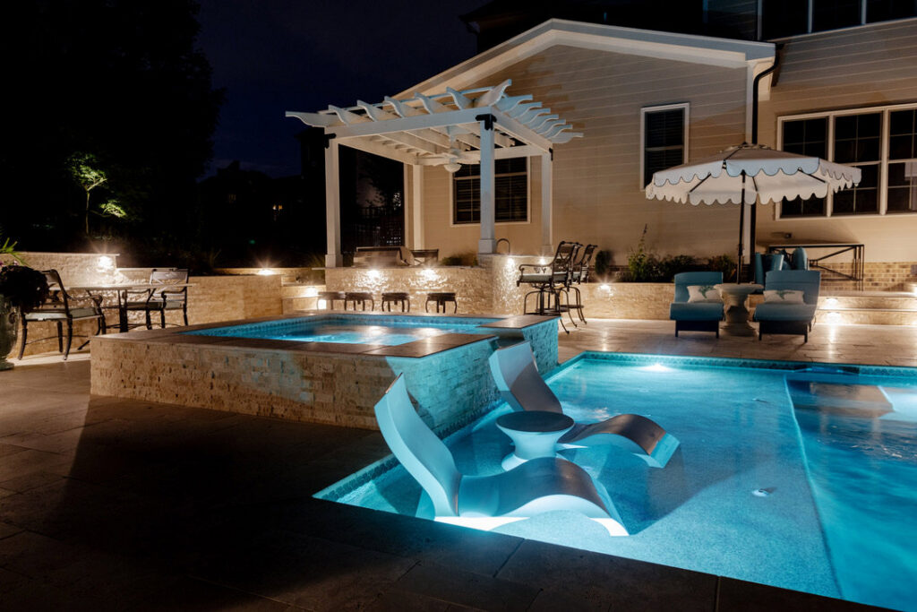 An illuminated night scene of a pool, hot tub, and pergola by Extreme Pools Renovation in Raleigh, NC.