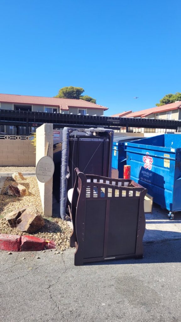 Old furniture and appliances, including a crib and treadmill, placed next to a dumpster for junk removal in Las Vegas, NV