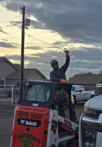 An operator in a branded Bobcat S70 skid-steer loader from ALL around junk, LCC, giving a peace sign in Mesa, AZ.