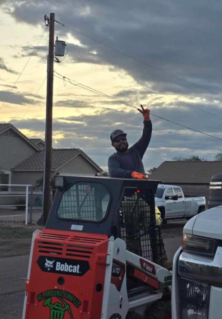 An operator in a branded Bobcat S70 skid-steer loader from ALL around junk, LCC, giving a peace sign in Mesa, AZ.